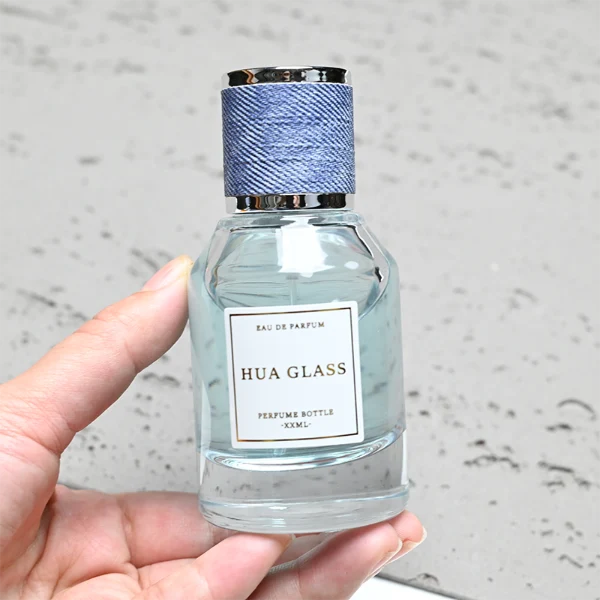 HUA0365 perfume bottle, cylindrical clear glass body with faceted sloped shoulders, silver neck and a paper label attached, textured blue or beige cylindrical plastic cap with silver trim, contemporary casual luxury fragrance packaging design available in 30ml, 50ml, and 100ml