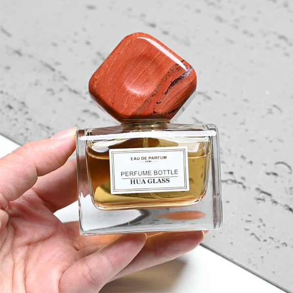 HUA0375 perfume bottle, wide rectangular cuboid clear glass body with centered paper label, oversized multi-faceted geometric semi-precious stone cap in jade green and reddish-brown tiger's eye gemstone textures, Zen luxury organic fragrance packaging design in 50ml