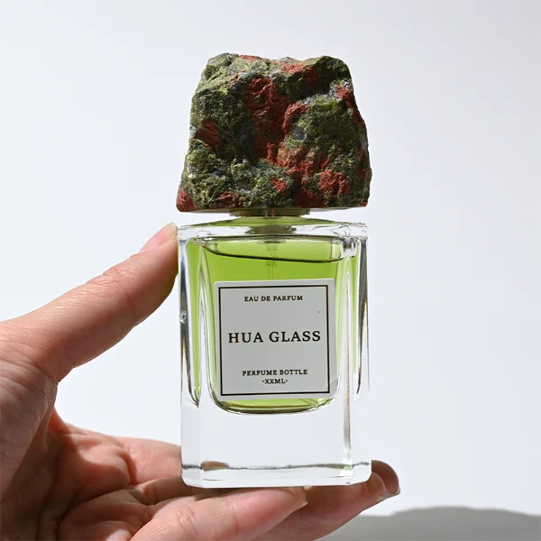 HUA0379 perfume bottle, cylindrical clear glass body with centered paper label, rugged organic raw semi-precious stone cap in pink rose quartz, tiger's eye jasper, green bloodstone or black lava rock textures, artistic luxury fragrance packaging design available in 30ml, 50ml, and 100ml