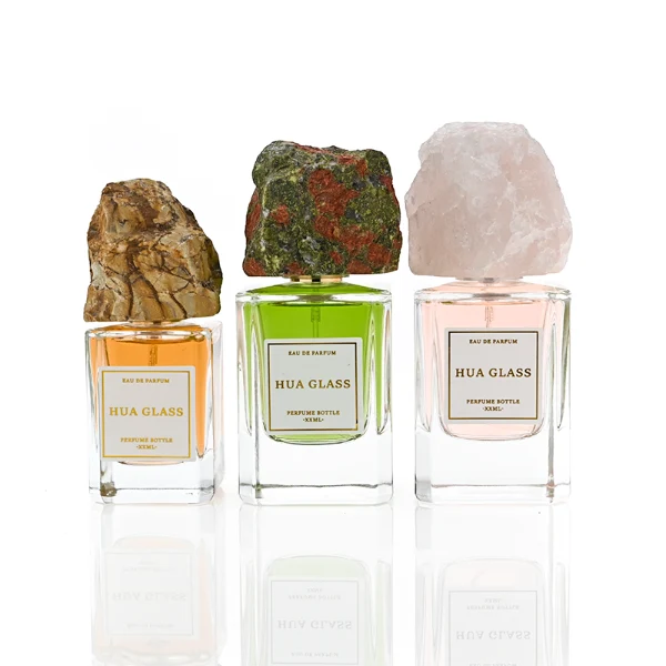 HUA0379 perfume bottle, cylindrical clear glass body with centered paper label, rugged organic raw semi-precious stone cap in pink rose quartz, tiger's eye jasper, green bloodstone or black lava rock textures, artistic luxury fragrance packaging design available in 30ml, 50ml, and 100ml
