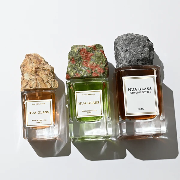 HUA0379 perfume bottle, cylindrical clear glass body with centered paper label, rugged organic raw semi-precious stone cap in pink rose quartz, tiger's eye jasper, green bloodstone or black lava rock textures, artistic luxury fragrance packaging design available in 30ml, 50ml, and 100ml