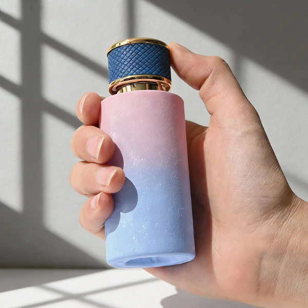 HUA0411 perfume bottle, luxury cylindrical glass bottle with glossy spray colored multi-color finish in pink or pink-blue gradient, featuring a gold-colored collar and a unique textured leather-wrapped plastic cap with gold-colored trim, modern elegant fragrance packaging design available in 30ml, 50ml, and 100ml