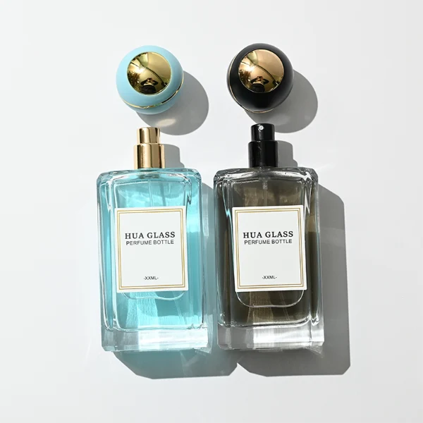 HUA0426 perfume bottle, luxury heavy-base rectangular square glass bottle with high-transparency finish showing colored liquid, featuring a gold-bordered square paper label and a unique round ball plastic cap with a diagonal gold-colored accent stripe, modern elegant fragrance packaging design available in 30ml, 50ml, and 100ml