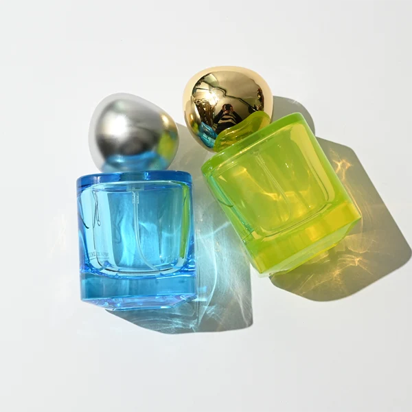 HUA0429 perfume bottle, luxury heavy-base round cylindrical glass bottle with high-transparency colorful finish showing blue, green, or brown liquid, featuring a unique irregular pebble-shaped plastic cap with a mirror-shine metallic chrome finish, modern artistic fragrance packaging design available in 30ml, 50ml, and 100ml