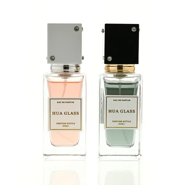 Two tall rectangular clear glass perfume bottles stand side by side, filled with light pink and grey-green liquids. Both feature a white square label and are topped with tall cube caps accented by tiny corner rhinestones—presented in white with a silver collar on the left, and black with a gold collar on the right.