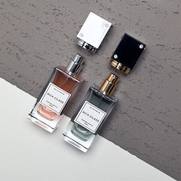 Two tall rectangular clear glass perfume bottles stand side by side, filled with light pink and grey-green liquids. Both feature a white square label and are topped with tall cube caps accented by tiny corner rhinestones—presented in white with a silver collar on the left, and black with a gold collar on the right.