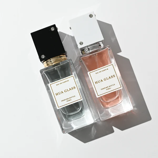 Two tall rectangular clear glass perfume bottles stand side by side, filled with light pink and grey-green liquids. Both feature a white square label and are topped with tall cube caps accented by tiny corner rhinestones—presented in white with a silver collar on the left, and black with a gold collar on the right.