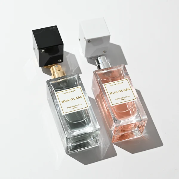 Two tall rectangular clear glass perfume bottles stand side by side, filled with light pink and grey-green liquids. Both feature a white square label and are topped with tall cube caps accented by tiny corner rhinestones—presented in white with a silver collar on the left, and black with a gold collar on the right.