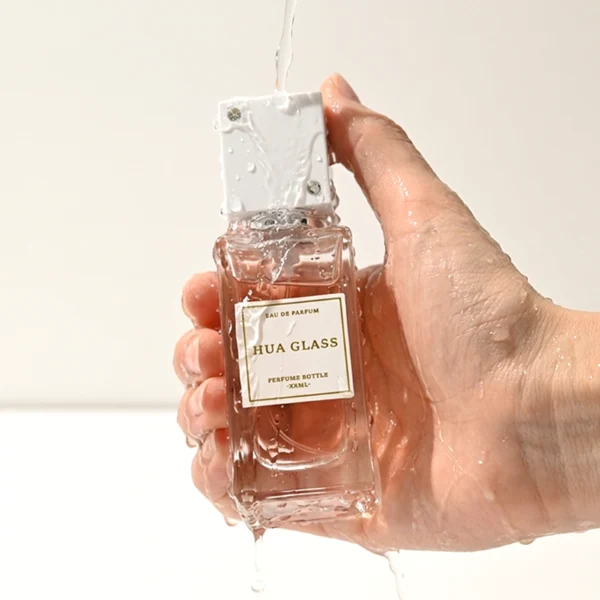 Two tall rectangular clear glass perfume bottles stand side by side, filled with light pink and grey-green liquids. Both feature a white square label and are topped with tall cube caps accented by tiny corner rhinestones—presented in white with a silver collar on the left, and black with a gold collar on the right.