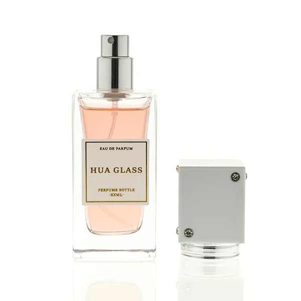 Two tall rectangular clear glass perfume bottles stand side by side, filled with light pink and grey-green liquids. Both feature a white square label and are topped with tall cube caps accented by tiny corner rhinestones—presented in white with a silver collar on the left, and black with a gold collar on the right.