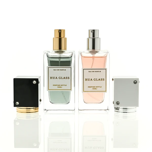 Two tall rectangular clear glass perfume bottles stand side by side, filled with light pink and grey-green liquids. Both feature a white square label and are topped with tall cube caps accented by tiny corner rhinestones—presented in white with a silver collar on the left, and black with a gold collar on the right.
