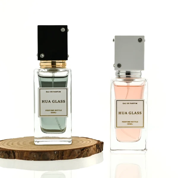 Two tall rectangular clear glass perfume bottles stand side by side, filled with light pink and grey-green liquids. Both feature a white square label and are topped with tall cube caps accented by tiny corner rhinestones—presented in white with a silver collar on the left, and black with a gold collar on the right.