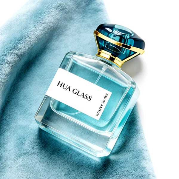 HUA0449 perfume bottle features a rounded short, thick-bottom square clear glass body paired with a unique organic, abstract-shaped translucent cap with a gold collar. Designed for wholesalers, it offers a 100ml capacity option, blending a modern minimalist profile with structural durability for distinct fragrance collections.