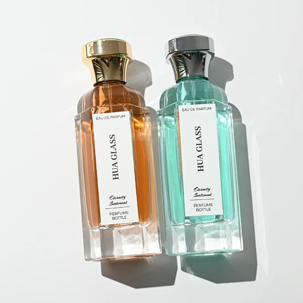 two tall clear hexagonal glass perfume bottles hold teal and amber liquids. The left bottle has a glossy silver cap, while the right bottle is topped with a polished gold cap.