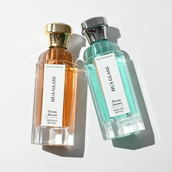 two tall clear hexagonal glass perfume bottles hold teal and amber liquids. The left bottle has a glossy silver cap, while the right bottle is topped with a polished gold cap.