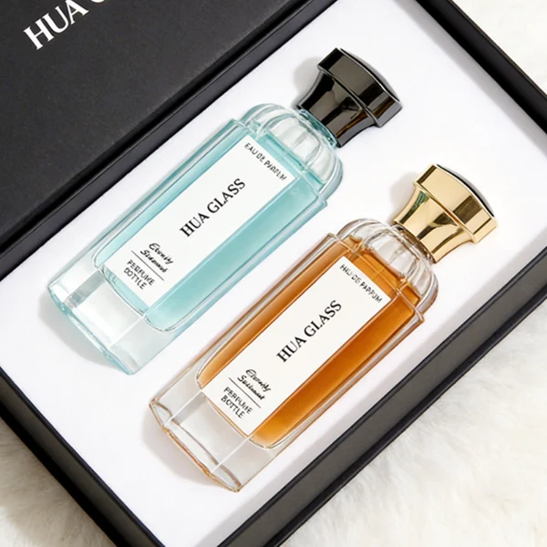 two tall clear hexagonal glass perfume bottles hold teal and amber liquids. The left bottle has a glossy silver cap, while the right bottle is topped with a polished gold cap.