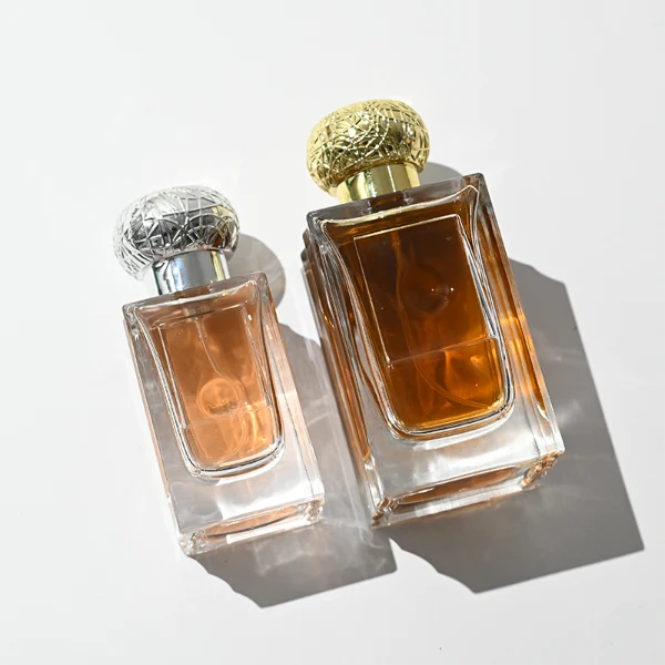 HUA0454 perfume bottle showcases a clear, thick-walled rectangular glass body with an ornate, textured round plastic cap in gold or silver. Designed for wholesalers, it offers 50ml and 100ml capacities, combining a sleek angular profile with a highly decorative cap style.