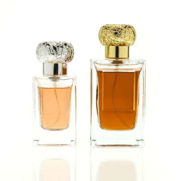 HUA0454 perfume bottle showcases a clear, thick-walled rectangular glass body with an ornate, textured round plastic cap in gold or silver. Designed for wholesalers, it offers 50ml and 100ml capacities, combining a sleek angular profile with a highly decorative cap style.