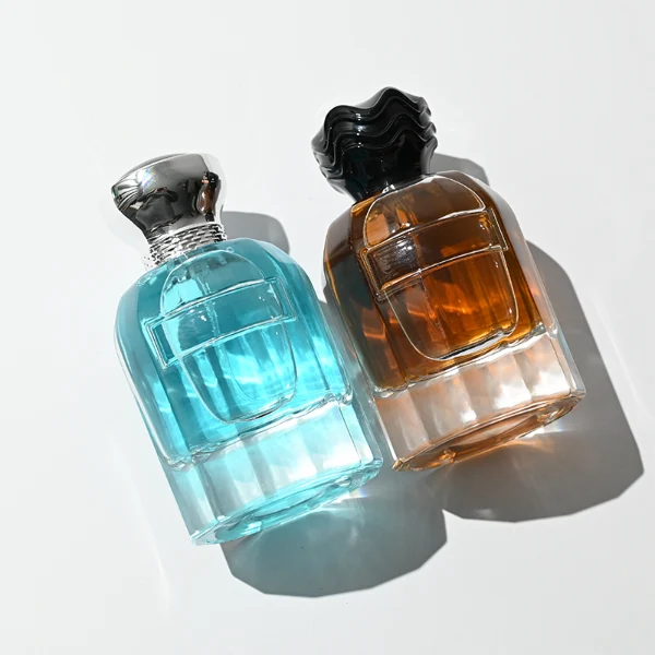 In the provided image, two clear Cylindrical glass perfume bottles with thick bases stand side by side, containing light blue and amber liquids. The left bottle is topped with a smooth, flared silver cap with a textured collar, while the right bottle is topped with a unique, textured wavy black cap.