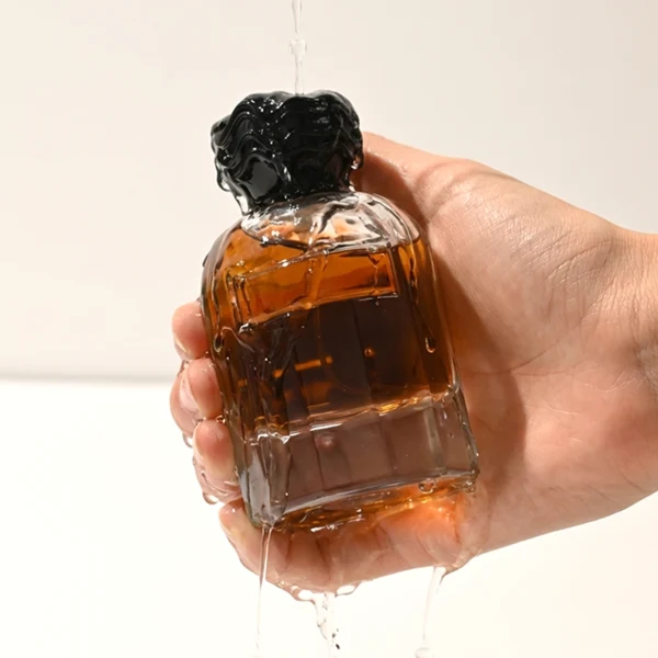 In the provided image, two clear Cylindrical glass perfume bottles with thick bases stand side by side, containing light blue and amber liquids. The left bottle is topped with a smooth, flared silver cap with a textured collar, while the right bottle is topped with a unique, textured wavy black cap.