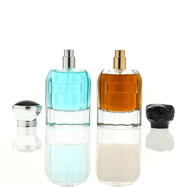In the provided image, two clear Cylindrical glass perfume bottles with thick bases stand side by side, containing light blue and amber liquids. The left bottle is topped with a smooth, flared silver cap with a textured collar, while the right bottle is topped with a unique, textured wavy black cap.