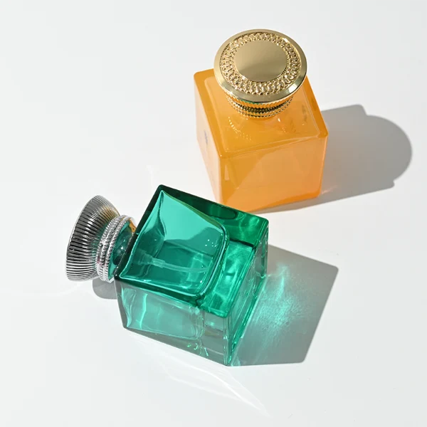 A thick-bottomed cube glass perfume bottle featuring an angular, square silhouette with semi-translucent or frosted colored finishes, topped with a heavy, uniquely ribbed metallic Zamac cap available in black, gold, or silver.