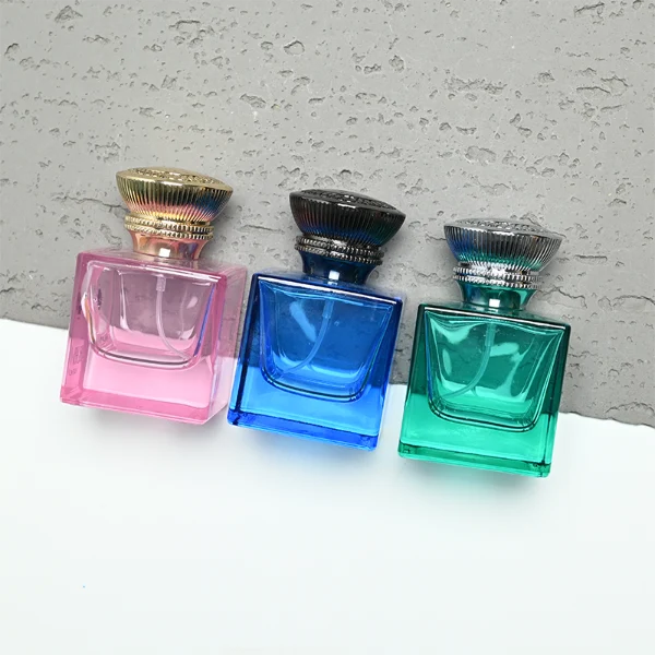 A thick-bottomed cube glass perfume bottle featuring an angular, square silhouette with semi-translucent or frosted colored finishes, topped with a heavy, uniquely ribbed metallic Zamac cap available in black, gold, or silver.
