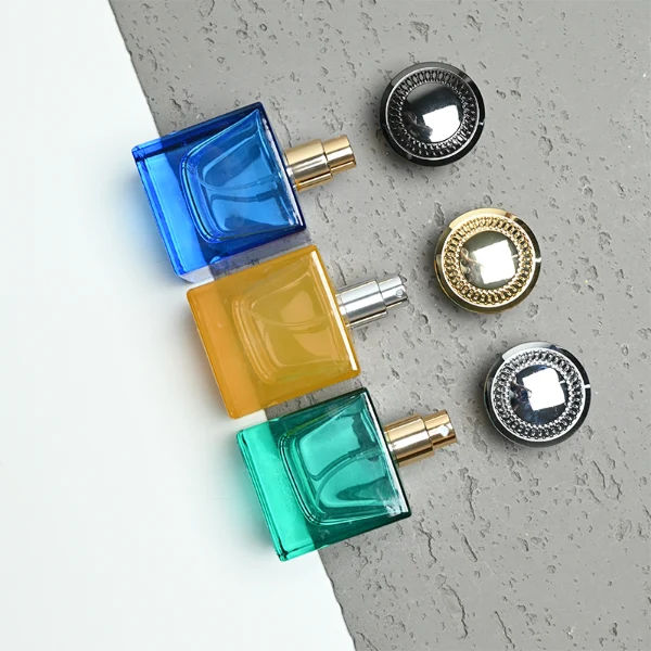 A thick-bottomed cube glass perfume bottle featuring an angular, square silhouette with semi-translucent or frosted colored finishes, topped with a heavy, uniquely ribbed metallic Zamac cap available in black, gold, or silver.
