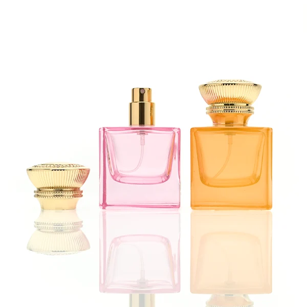 A thick-bottomed cube glass perfume bottle featuring an angular, square silhouette with semi-translucent or frosted colored finishes, topped with a heavy, uniquely ribbed metallic Zamac cap available in black, gold, or silver.