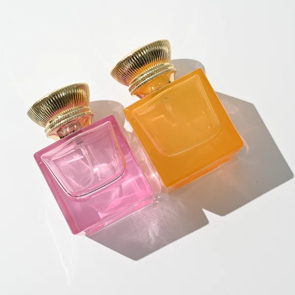 A thick-bottomed cube glass perfume bottle featuring an angular, square silhouette with semi-translucent or frosted colored finishes, topped with a heavy, uniquely ribbed metallic Zamac cap available in black, gold, or silver.