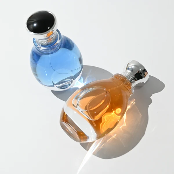 A 100ml clear glass perfume bottle with a smooth, globular curved profile and a thick base. The neck features a detailed metallic collar paired with a silver crown-motif cap topped with a dark glossy insert.