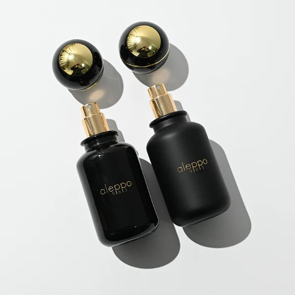 two cylindrical solid black perfume bottles stand side by side, one in a glossy finish and the other in a matte finish. Both bottles feature a gold logo that reads "aleppo 91613" and are topped with unique, spherical black caps encircled by a diagonal gold ribbon accent and a gold collar.