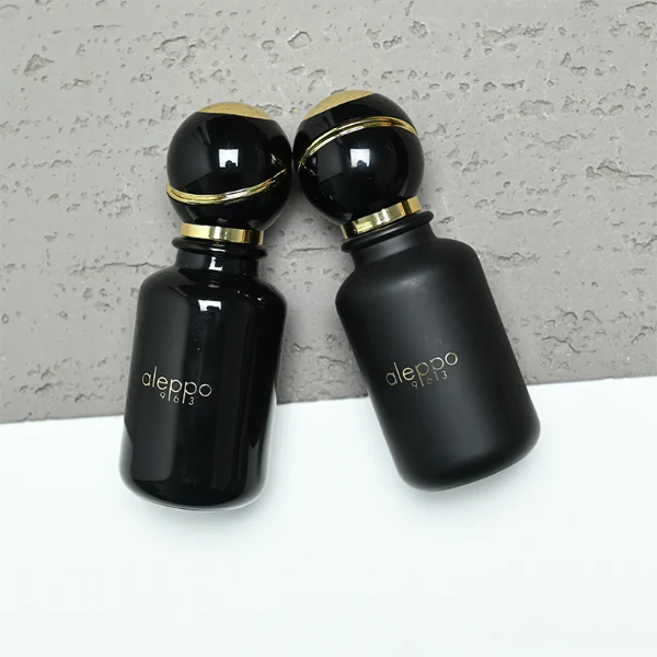two cylindrical solid black perfume bottles stand side by side, one in a glossy finish and the other in a matte finish. Both bottles feature a gold logo that reads "aleppo 91613" and are topped with unique, spherical black caps encircled by a diagonal gold ribbon accent and a gold collar.