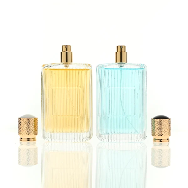 A rectangular clear glass perfume bottle detailed with distinct vertical grooves and a smooth central label indentation, paired with a cylindrical golden plastic cap featuring an intricate textured pattern.