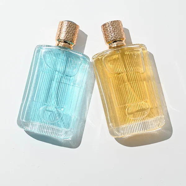 A rectangular clear glass perfume bottle detailed with distinct vertical grooves and a smooth central label indentation, paired with a cylindrical golden plastic cap featuring an intricate textured pattern.