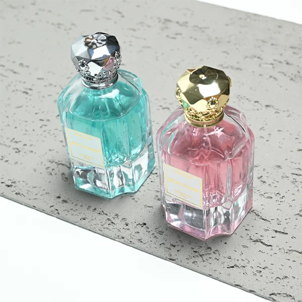 A hexagonal clear glass perfume bottle with structured vertical facets along the body, paired with an ornate, crown-shaped heavy metallic Zamac cap available in polished gold or silver.