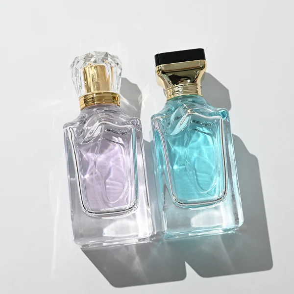 A thick-bottomed rectangular clear glass perfume bottle with faceted upper corners, shown with two cap variations: a crystal-clear faceted crown-shaped cap with a gold core, and a flared gold cap topped with a flat black cube block.