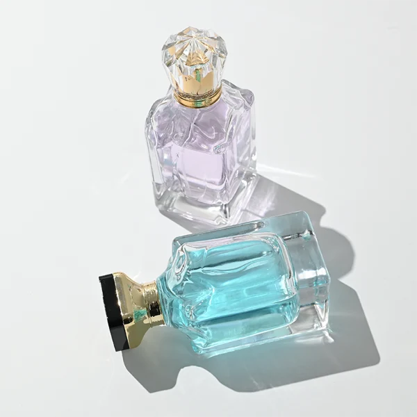 A thick-bottomed rectangular clear glass perfume bottle with faceted upper corners, shown with two cap variations: a crystal-clear faceted crown-shaped cap with a gold core, and a flared gold cap topped with a flat black cube block.