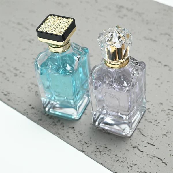 A thick-bottomed rectangular clear glass perfume bottle with faceted upper corners, shown with two cap variations: a crystal-clear faceted crown-shaped cap with a gold core, and a flared gold cap topped with a flat black cube block.