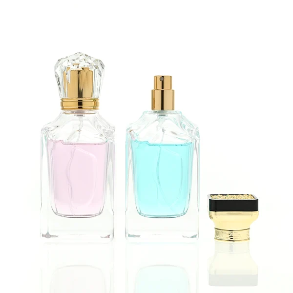 A thick-bottomed rectangular clear glass perfume bottle with faceted upper corners, shown with two cap variations: a crystal-clear faceted crown-shaped cap with a gold core, and a flared gold cap topped with a flat black cube block.