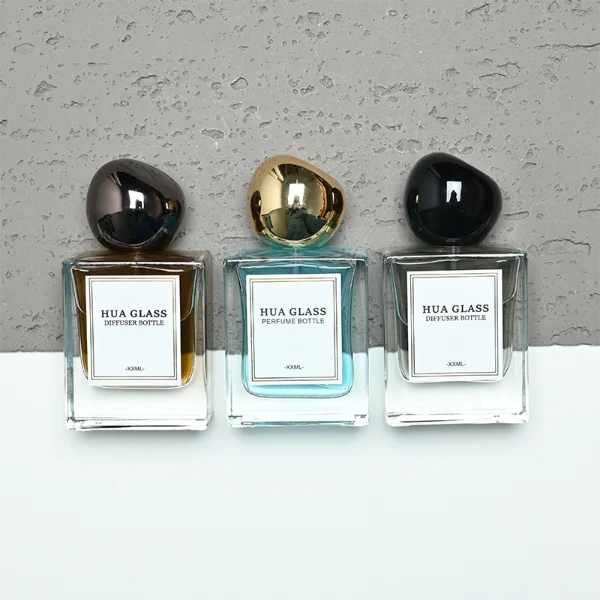 In the image, a thick-bottomed cube clear glass perfume bottle with a structured square silhouette is paired with a smooth, asymmetrical oval ellipse plastic cap available in polished gold, glossy black, or metallic brown finish.