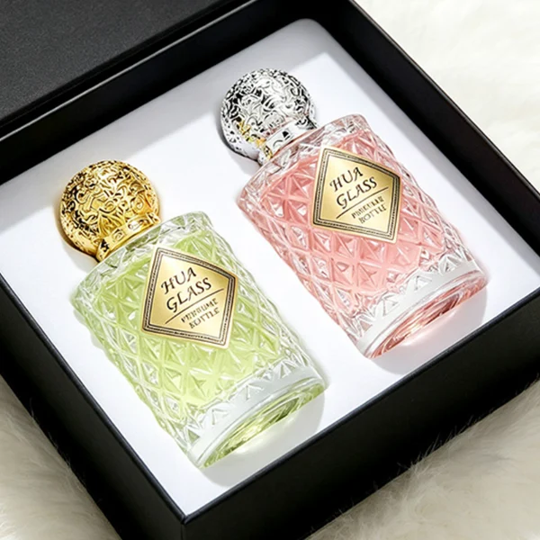 two clear textured glass perfume bottles hold light green and pink liquids. The bottles are topped with round, ornate filigree caps finished in polished gold on the left and bright silver on the right.