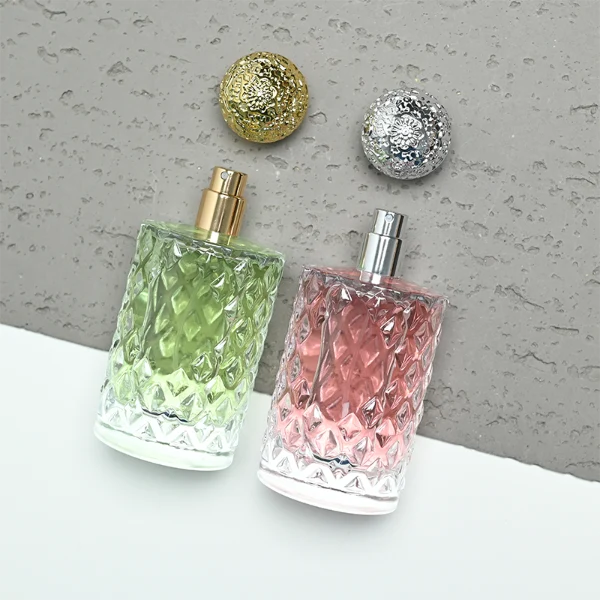 two clear textured glass perfume bottles hold light green and pink liquids. The bottles are topped with round, ornate filigree caps finished in polished gold on the left and bright silver on the right.