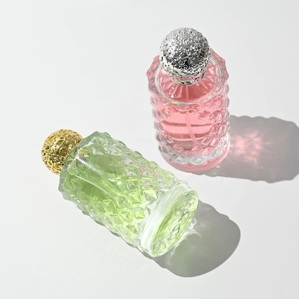 two clear textured glass perfume bottles hold light green and pink liquids. The bottles are topped with round, ornate filigree caps finished in polished gold on the left and bright silver on the right.