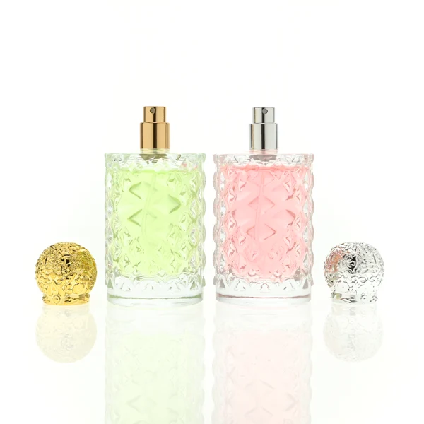 two clear textured glass perfume bottles hold light green and pink liquids. The bottles are topped with round, ornate filigree caps finished in polished gold on the left and bright silver on the right.