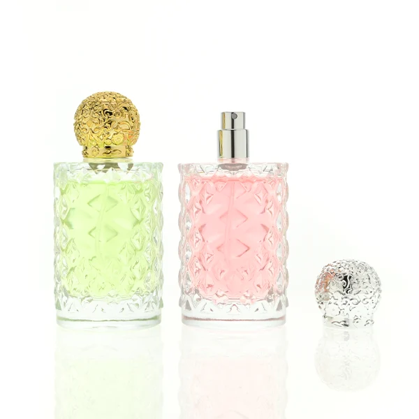 two clear textured glass perfume bottles hold light green and pink liquids. The bottles are topped with round, ornate filigree caps finished in polished gold on the left and bright silver on the right.