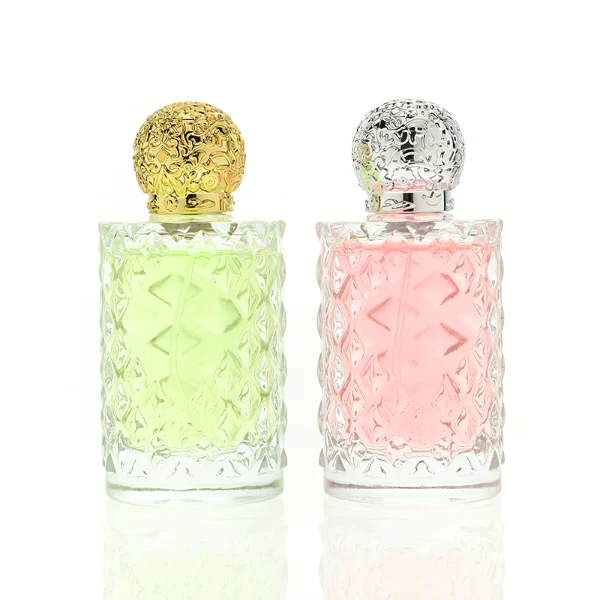 two clear textured glass perfume bottles hold light green and pink liquids. The bottles are topped with round, ornate filigree caps finished in polished gold on the left and bright silver on the right.