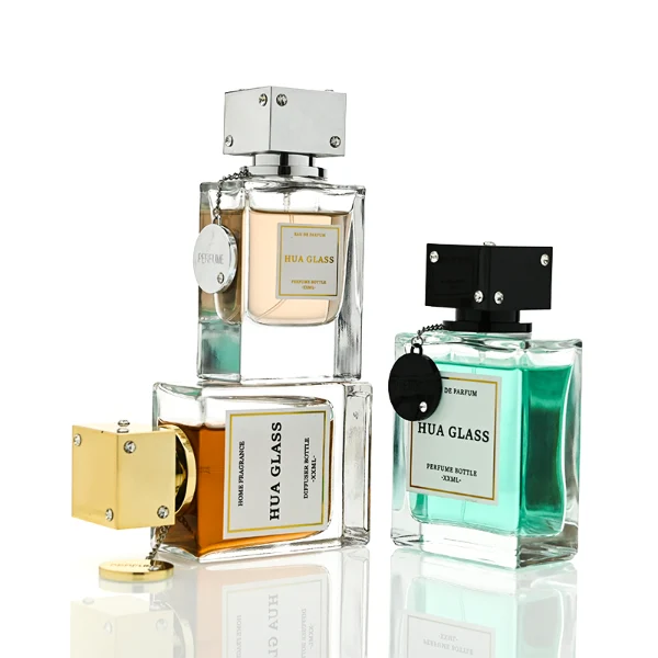 A lineup of three thick-bottomed rectangular clear glass perfume bottles containing peach, amber, and light green liquids. Each bottle is topped with a distinct cube-shaped cap accented with small rhinestone studs at the corners and a delicate metal hanging chain with a circular tag attached to the neck. The caps are finished in polished metallic silver, polished metallic gold, and solid black.