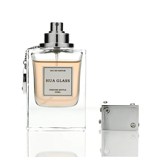 A lineup of three thick-bottomed rectangular clear glass perfume bottles containing peach, amber, and light green liquids. Each bottle is topped with a distinct cube-shaped cap accented with small rhinestone studs at the corners and a delicate metal hanging chain with a circular tag attached to the neck. The caps are finished in polished metallic silver, polished metallic gold, and solid black.