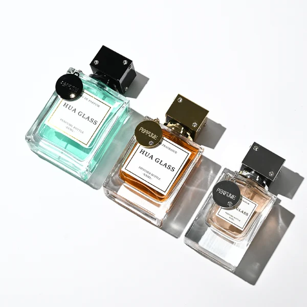 A lineup of three thick-bottomed rectangular clear glass perfume bottles containing peach, amber, and light green liquids. Each bottle is topped with a distinct cube-shaped cap accented with small rhinestone studs at the corners and a delicate metal hanging chain with a circular tag attached to the neck. The caps are finished in polished metallic silver, polished metallic gold, and solid black.